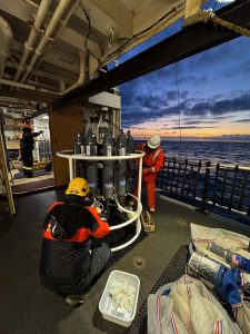 Two crew members on the deck of a ship make adjustments to a machine with many tanks and tubes. 
