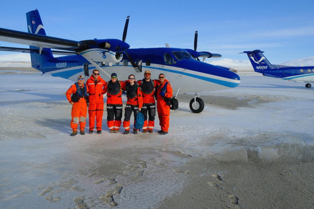 A group of researchers dressed in jumpsuits poses in front of a NOAA Twin Otter airplane.