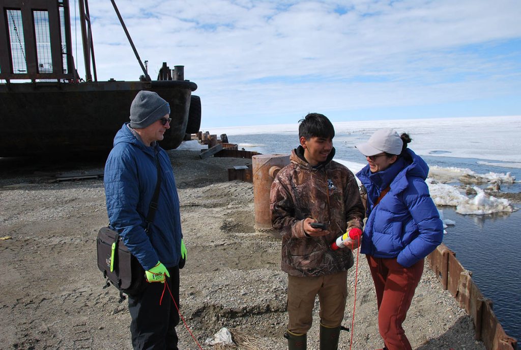 Three researchers in winter clothing stand near the shore on a bright, cold day.