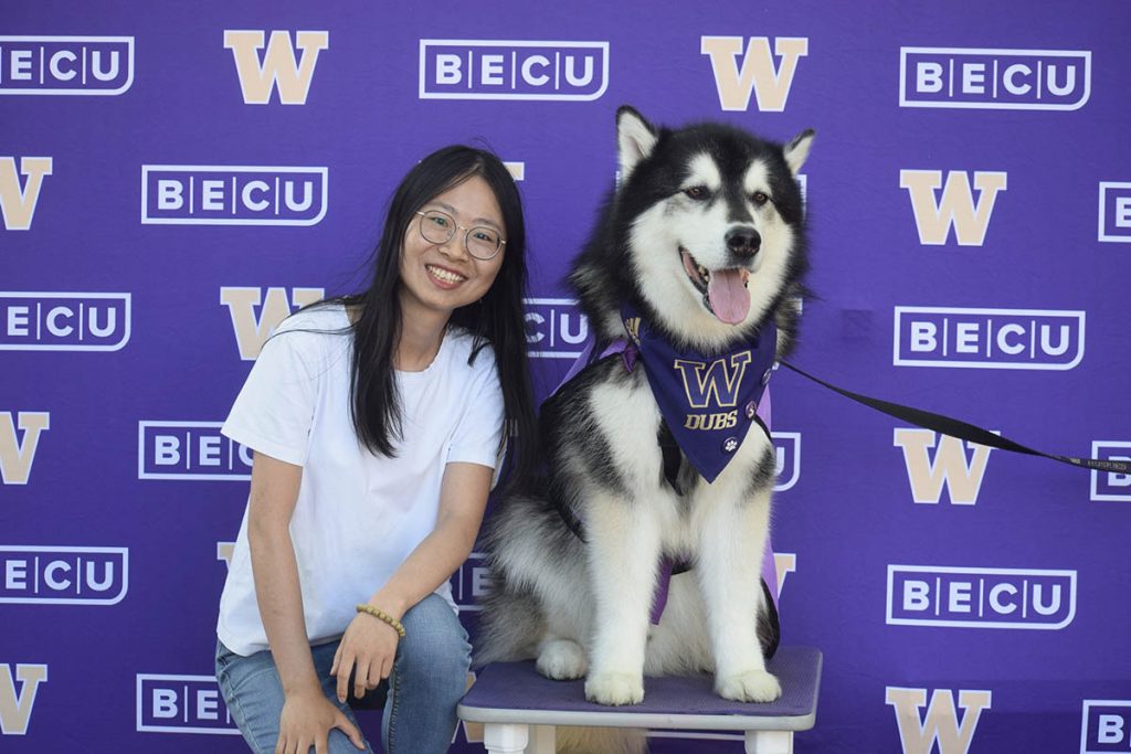 Qiuxian Li poses with a husky in front of a University of Washington backdrop.