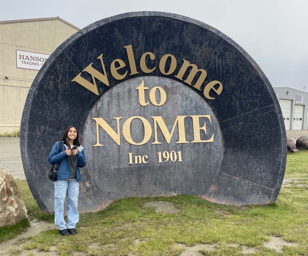An intern poses next to a large gold-pan-shaped sign that says "Welcome to Nome".