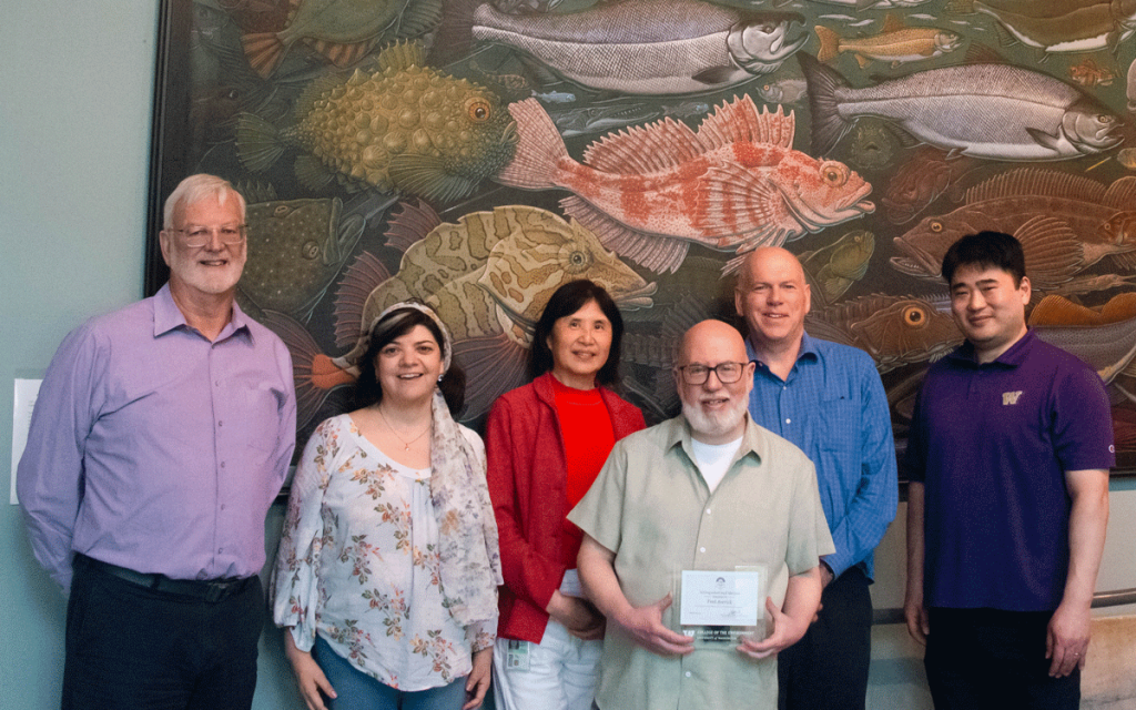 A small group of CICOES employees gather around Fred, who is holding an award. 