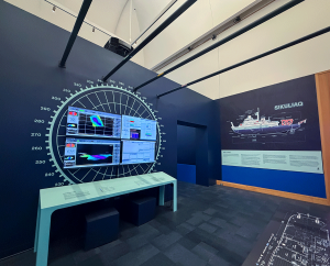 A display at the Anchorage Museum, showing John Horne's video alongside others, as well as a diagram of the Sikuliaq in the distance. 