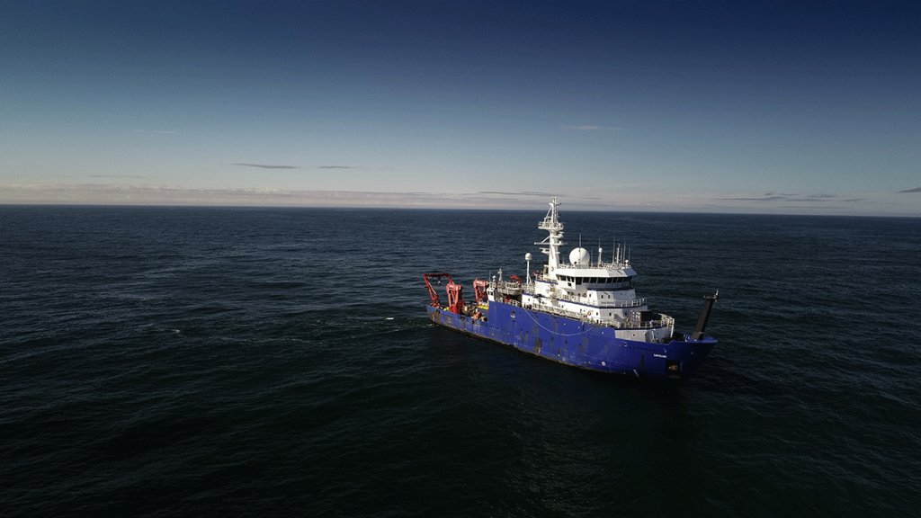 An aerial view of the R/V Sikuliaq at sea, with the sun low on the horizon.