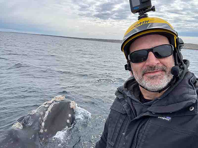 A selfie of Alex Zerbini on a small boat, with a humpback whale in the background.