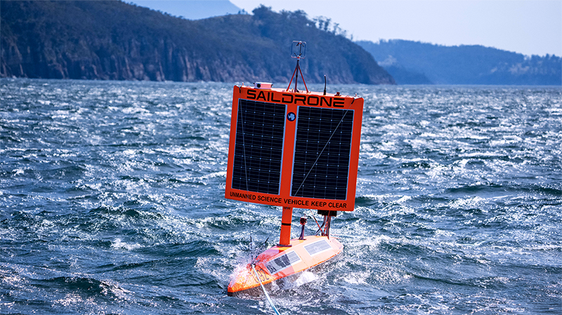 A sturdy, bright orange water vessel called a Saildrone is towed through choppy water.